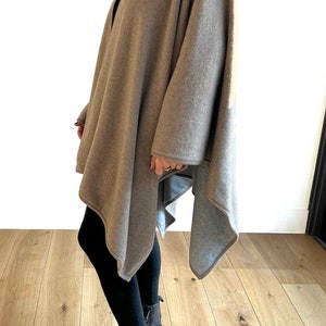 May include: A brown knitted poncho with a long, asymmetrical hemline. The poncho is worn over black leggings and black boots.