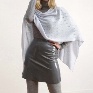 May include: A woman wearing a light blue knitted poncho over a gray turtleneck sweater and a gray skirt. She is standing on a wooden floor.