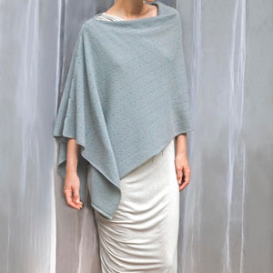 May include: A light blue knitted poncho with a delicate pattern. The poncho is draped over a white dress.