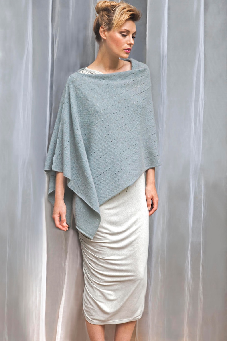 May include: A light blue knitted poncho with a delicate pattern. The poncho is draped over a white dress.