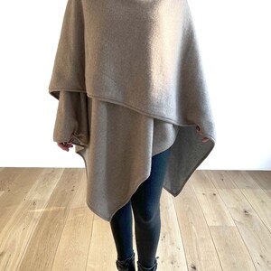 May include: A beige cashmere wrap with a draped design. The wrap is worn over black leggings and black boots.