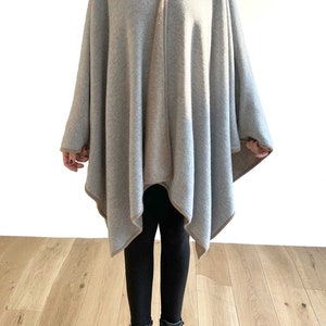 May include: A gray and beige knitted poncho with a triangular shape. The poncho is worn over black leggings and black boots.