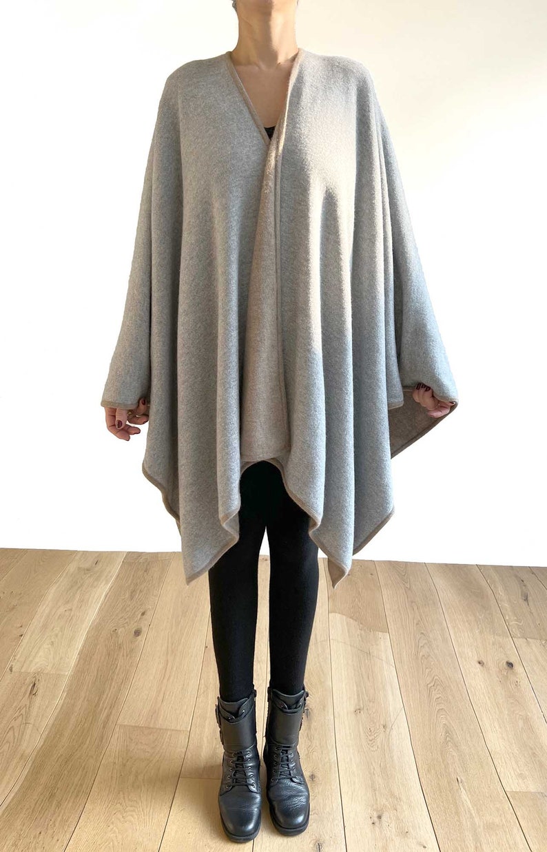 May include: A gray and beige knitted poncho with a triangular shape. The poncho is worn over black leggings and black boots.