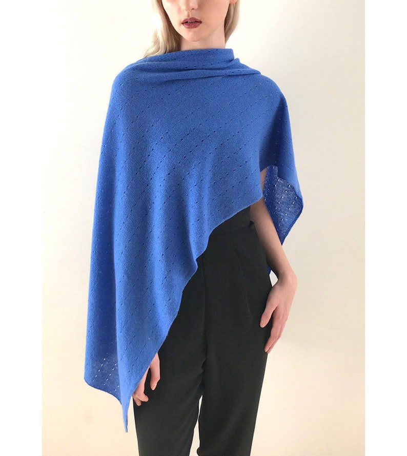 May include: A blue knitted poncho with a delicate openwork pattern. The poncho is draped over a person's shoulders and is styled with black pants.