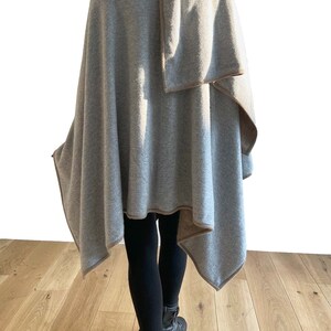 May include: A gray cashmere wrap with a brown trim. The wrap is folded over the shoulder and draped down the back. The person is wearing black leggings and black boots.