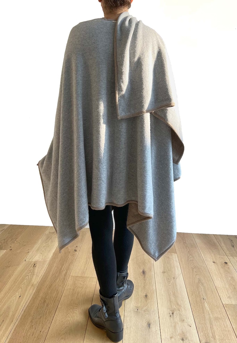 May include: A gray cashmere wrap with a brown trim. The wrap is folded over the shoulder and draped down the back. The person is wearing black leggings and black boots.