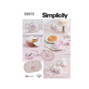 May include: Simplicity pattern S9573 featuring a variety of tea party accessories. The image showcases a teapot, teacups, a basket, placemats, and a tea set. The items are in pastel colors with floral and paisley designs. The pattern is designed and printed in the USA.