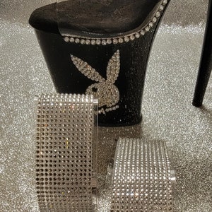 May include: A pair of black platform heels with a clear strap and a rhinestone-covered design. The heels feature a Playboy bunny logo and are adorned with rhinestones. Two additional rhinestone-covered bands are also included.