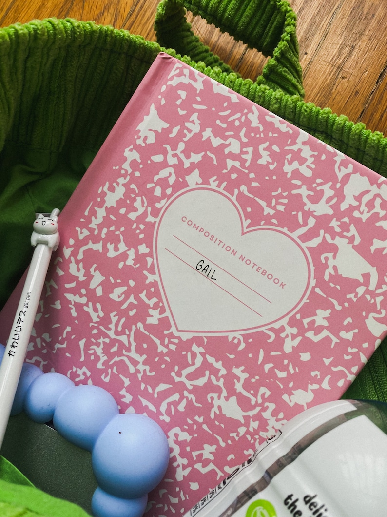 May include: Pink composition notebook with a white heart and the word "GAIL" written inside. The notebook is in a green bag with a blue and white toy.