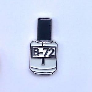 May include: White enamel pin of a bottle of nail polish with a black cap and a label that says "B-72". The pin is on a white card with the text "Preservation Pins from Hangerbee" and "www.hangerbee.com".