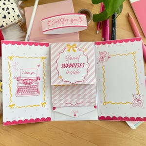 Può includere: Un biglietto di auguri rosa e bianco con la scritta "Sweet Surprises Inside" e "I love you". Il biglietto presenta un bordo decorativo e illustrazioni di una macchina da scrivere, un orsacchiotto e un angelo. Sono visibili anche un cuore rosso all'uncinetto e articoli di cancelleria rosa.