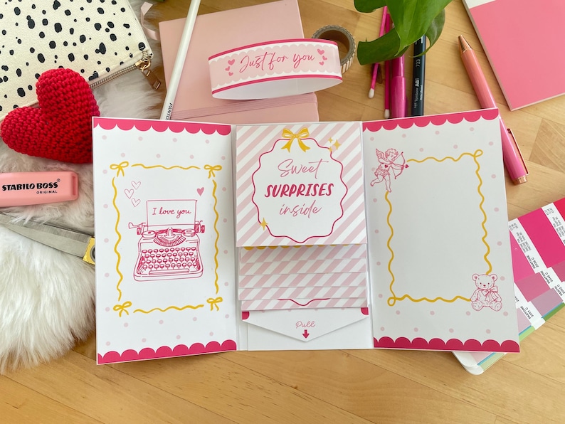 以下が含まれることがあります： 「Sweet Surprises Inside」と「I love you」の文字が入ったピンクと白のグリーティングカード。カードには装飾的なボーダーと、タイプライター、テディベア、天使のイラストが描かれています。赤いかぎ針編みのハートとピンクの文房具も見えます。