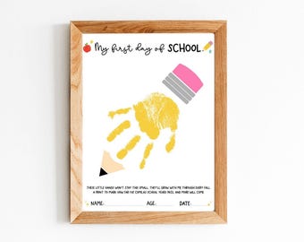 May include: A framed print with the text "My first day of SCHOOL." features a yellow handprint and a pencil illustration. The print includes space to record the child's name, age, and date. The frame is made of light brown wood.