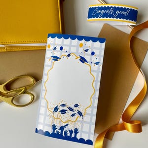 May include: A graduation card with a blue and gold design, featuring graduation caps and hands reaching upwards. A gold ribbon, scissors, and a yellow notebook are also visible. The card has a blank space for a photo.