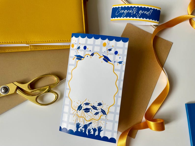 May include: A graduation card with a blue and gold design, featuring graduation caps and hands reaching upwards. A gold ribbon, scissors, and a yellow notebook are also visible. The card has a blank space for a photo.