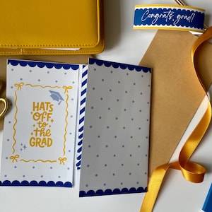 May include: A graduation-themed flat lay with a yellow wallet, gold scissors, a card with the text "Hats Off to the Grad", and a ribbon. The card has a blue scalloped border and a graduation cap illustration.