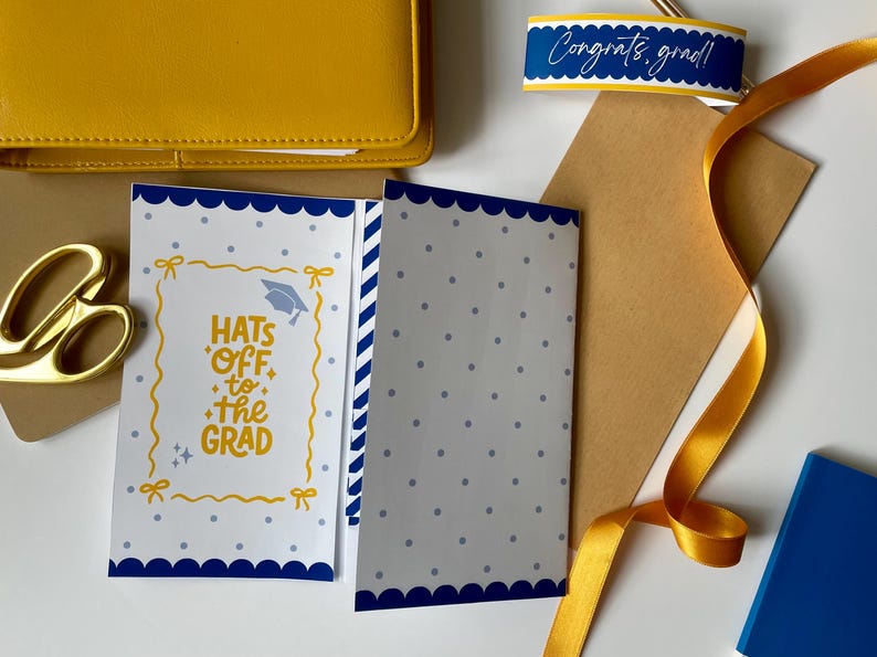 May include: A graduation-themed flat lay with a yellow wallet, gold scissors, a card with the text "Hats Off to the Grad", and a ribbon. The card has a blue scalloped border and a graduation cap illustration.