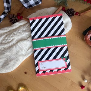 May include: A holiday gift card with a black and white striped pattern, scalloped red edges, and a green band. The card has a space to write "To:" and "From:" and is decorated with holly.