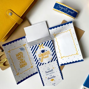 May include: A graduation-themed stationery set featuring a yellow notebook, scissors, and cards with blue and white designs. The cards have text that reads "Congrats Grad" and "Hats off to the Grad."