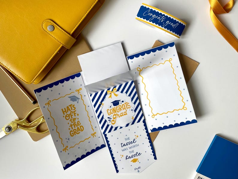 May include: A graduation-themed stationery set featuring a yellow notebook, scissors, and cards with blue and white designs. The cards have text that reads "Congrats Grad" and "Hats off to the Grad."