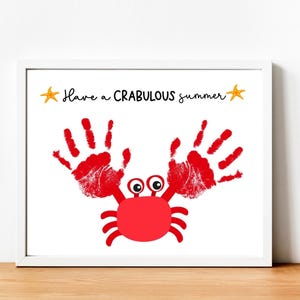 Puede incluir: Una impresión enmarcada blanca con el texto "Have a CRABULOUS summer" y dos estrellas de mar amarillas. Un cangrejo rojo hecho con huellas de manos está en el centro de la impresión.