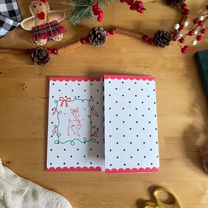 May include: Open Christmas card with a white background and black polka dots. The left side features a red and green illustrated reindeer design. The card has a scalloped pink border. Festive holiday decor surrounds the card.