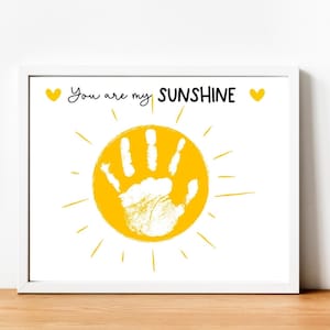 Puede incluir: Un marco blanco con un sol amarillo y una huella de mano blanca en su interior. El texto "You are my SUNSHINE" está escrito sobre el sol.