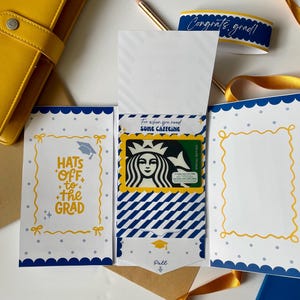 May include: A graduation gift card holder with a Starbucks gift card. The holder is white with blue and yellow accents, featuring the text "Hats off to the Grad" and "For when you need some caffeine". A yellow ribbon and a blue ribbon with the text "Congrats, grad!" are also present.