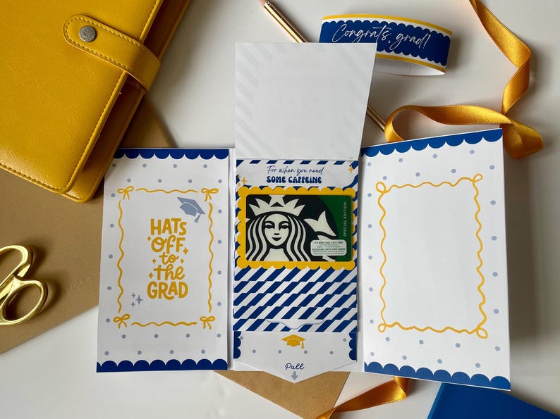 May include: A graduation gift card holder with a Starbucks gift card. The holder is white with blue and yellow accents, featuring the text "Hats off to the Grad" and "For when you need some caffeine". A yellow ribbon and a blue ribbon with the text "Congrats, grad!" are also present.