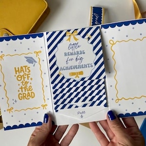 May include: A graduation card with a blue and white design. The card says "Hats off to the Grad" and "Little rewards for big achievements". The card has a pull tab and a yellow bow.