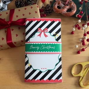 May include: A Christmas-themed notebook with a black and white striped pattern, red scalloped edges, and a green band that reads "Merry Christmas." The notebook has a red bow and star accents.