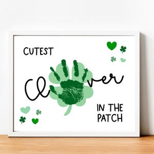 May include: White framed print with the text "Cutest Clover in the Patch". The artwork features a green handprint clover design with green hearts and shamrocks. The print is displayed on a wooden surface.