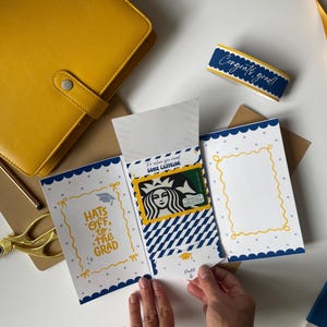 May include: A graduation gift set featuring a yellow leather journal, a card with the text "Hats off to the Grad", and a Starbucks gift card. The card has a blue and white scalloped border and a yellow frame.