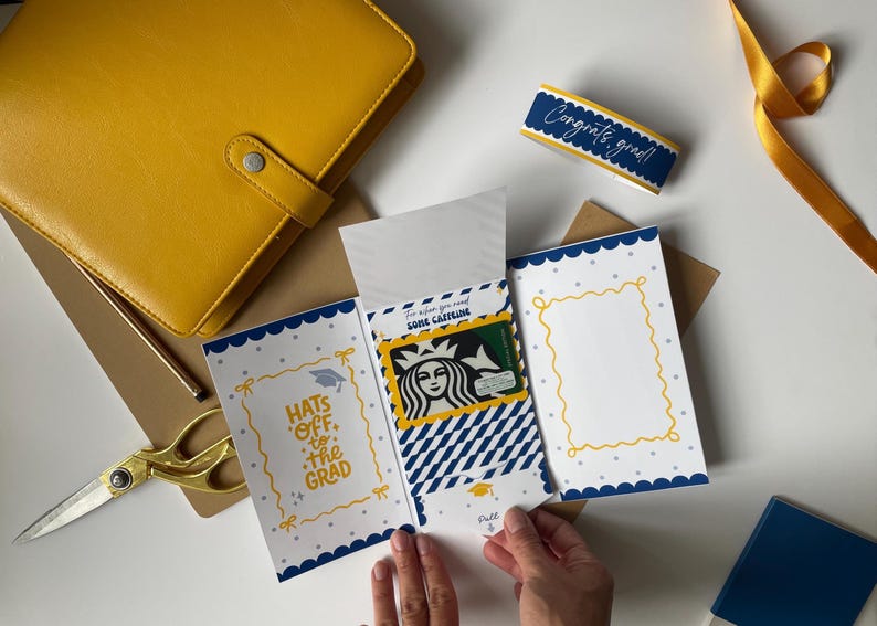 May include: A graduation gift set featuring a yellow leather journal, a card with the text "Hats off to the Grad", and a Starbucks gift card. The card has a blue and white scalloped border and a yellow frame.