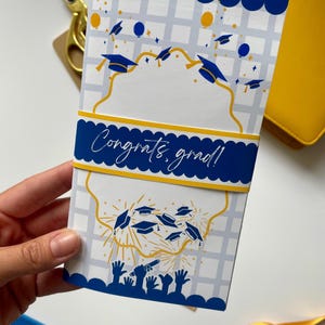 May include: A graduation card with a blue and white design. The card features a blue banner with the text "Congrats, grad!" and illustrations of graduation caps and fireworks. The card is held by a hand.
