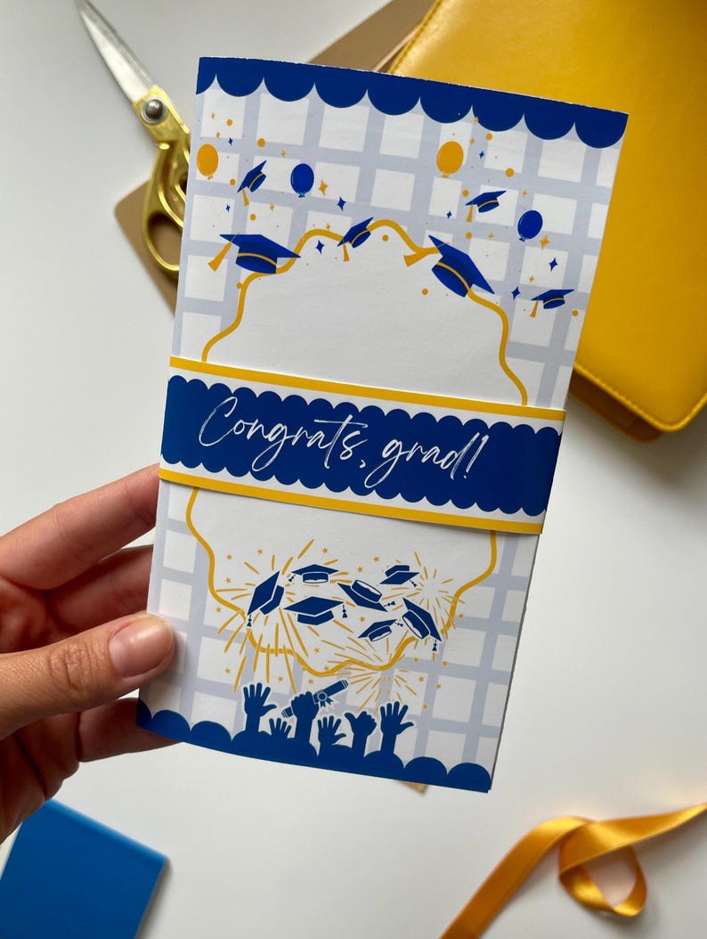 May include: A graduation card with a blue and white design. The card features a blue banner with the text "Congrats, grad!" and illustrations of graduation caps and fireworks. The card is held by a hand.