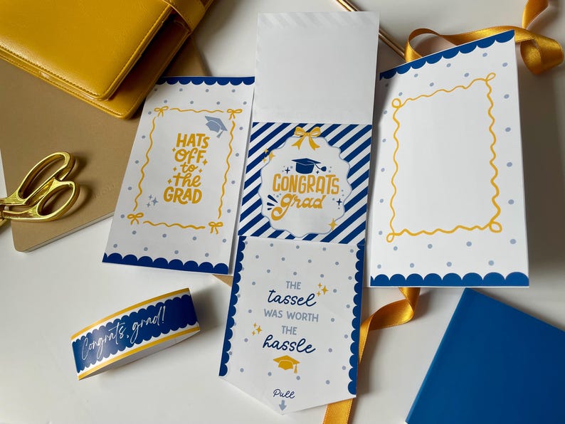 May include: A graduation stationery set in blue and yellow. The set includes cards with scalloped edges and text such as "Hats off to the Grad" and "Congrats Grad." A matching ribbon and a blue notebook are also visible.