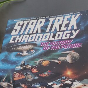 May include: A hardcover book titled "Star Trek Chronology: The History of the Future" is displayed on a clear stand. The cover features a space scene with the Star Trek logo and images of characters and spacecraft. The authors are Michael Okuda and Denise Okuda.