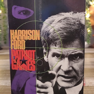 May include: A VHS tape box for the movie "Patriot Games" starring Harrison Ford. The box features a split design with a masked figure and Ford aiming a gun. Text includes the movie title and the tagline "Not for honor. Not for country. For his wife and child."