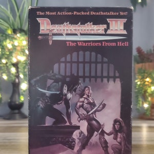 Puede incluir: Una cinta de VHS de Deathstalker III. La portada presenta una ilustración fantástica de guerreros musculosos con espadas, una mujer y el texto "The Warriors From Hell". La cinta tiene formato vertical.