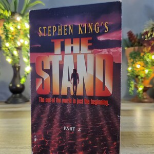 May include: A VHS tape of Stephen King's "The Stand, Part 2." The cover features a dark sky with the title in large orange letters. A silhouetted figure walks toward the viewer. The tagline reads, "The end of the world is just the beginning."