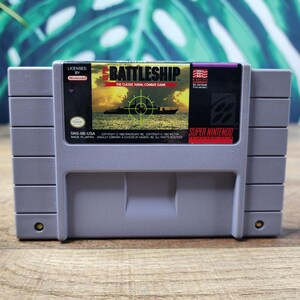 Puede incluir: Un cartucho de juego gris de Super Nintendo para el juego Battleship. La etiqueta presenta un atardecer amarillo sobre el océano con la silueta de un barco negro y una retícula de puntería verde. El texto dice BATTLESHIP THE CLASSIC NAVAL COMBAT GAME.