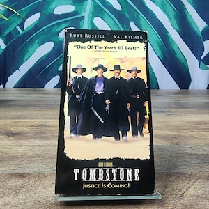 Peut inclure: Une cassette VHS du film "Tombstone" avec Kurt Russell et Val Kilmer. La jaquette montre quatre hommes en manteaux noirs et chapeaux. Le texte sur la cassette indique "One Of The Year's 10 Best!" et "Justice Is Coming!"