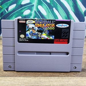 Puede incluir: Un cartucho de videojuego gris de Super Nintendo para el título Super Baseball Simulator 1.000. La etiqueta muestra a un jugador de béisbol con uniforme azul, el título del juego en texto amarillo y blanco, y los logotipos de Nintendo y Culture Brain.