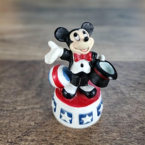May include: A small figurine of Mickey Mouse dressed in a tuxedo and red bow tie, holding a top hat. He stands on a red, white, and blue circus-style platform with star designs. A red, white, and blue ball is also present.