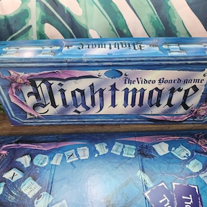 May include: A blue board game box and game board with the title "Nightmare". The box has a distressed wood-like appearance with purple bat-like creatures. The game board is blue with game pieces and text.