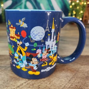 May include: A navy blue ceramic mug featuring a colorful illustration of Disney characters and landmarks. The design includes Mickey Mouse, Minnie Mouse, Goofy, and Dumbo, with the Eiffel Tower and Cinderella's castle. The mug has a large handle.