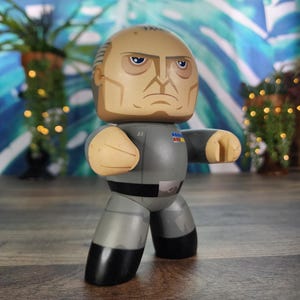 Puede incluir: Figura de acción estilizada de un personaje de rostro severo con uniforme gris y botas negras. La figura tiene una cabeza color canela con expresión seria y posa con los puños cerrados. El fondo presenta un patrón de hojas tropicales.