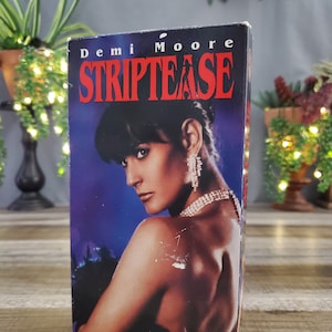 May include: A VHS tape for the movie "Striptease" starring Demi Moore. The cover features a portrait of Moore in a glamorous pose, with the title in bold red letters against a dark blue background. The tape is displayed on a wooden surface.