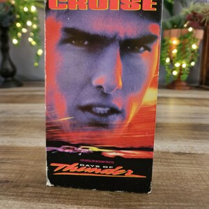 May include: A vintage VHS tape for the movie "Days of Thunder" featuring a close-up of a man's face with the name "CRUISE" at the top. The background has a red and orange gradient.